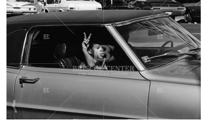 Photograph of a youthful passenger flashing a peace sign while driving in downtown Austin, circa 1965-1970
